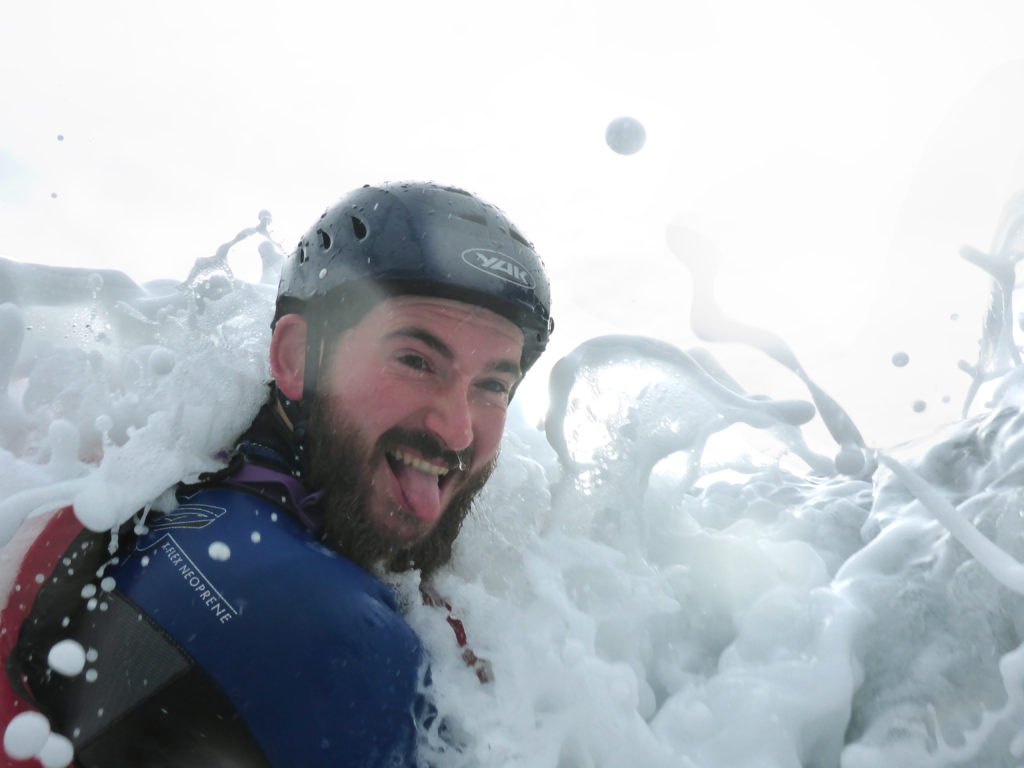 man looking at the camera whilst being splashed by a wave whilst coasteering